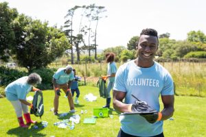 Portrait of a smiling young African American man standing in a field wearing wearing a t shirt with volunteer written on it and gloves, writing on a clipboard and smiling to camera, while a diverse group of volunteers collect rubbish and recycling in the