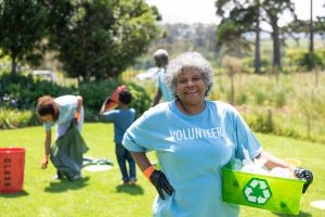 Front view of a senior African American woman standing in a field wearing a t shirt with volunteer written on it, holding a green plastic recycling crate and smiling to camera, with a diverse group of adults and children collecting rubbish and recycling i